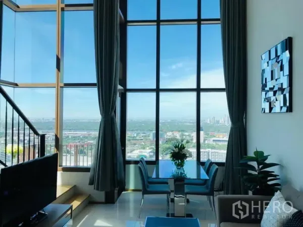 Condominium for rent in Khlong Toei, Bangkok - Bright living-dining space with panoramic skyline through tall windows and a staircase in a Khlong Toei condominium.
