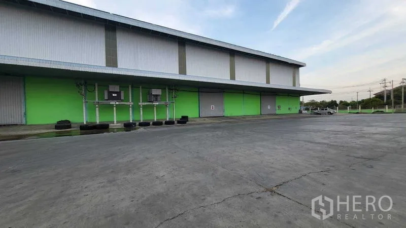 Factory for rent in Sai Noi, Nonthaburi - Side facade with green walls, several roller shutter doors, utilities and a wide apron.