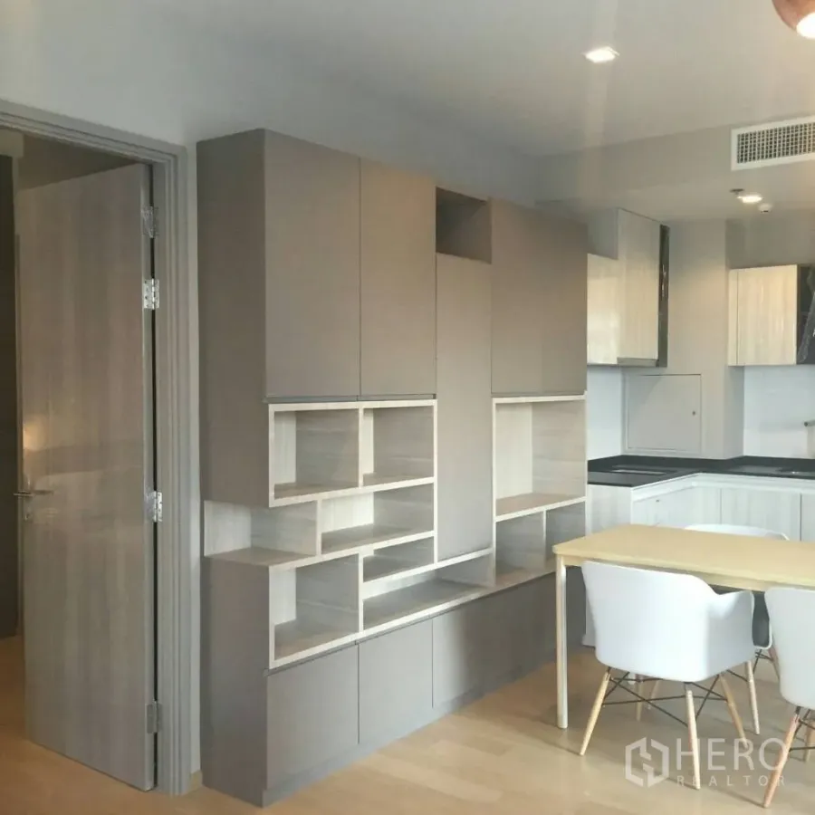 Condominium for rent in Watthana, Bangkok - Built-in storage wall with open shelves beside a compact dining table and L-shaped kitchen in a modern condo.