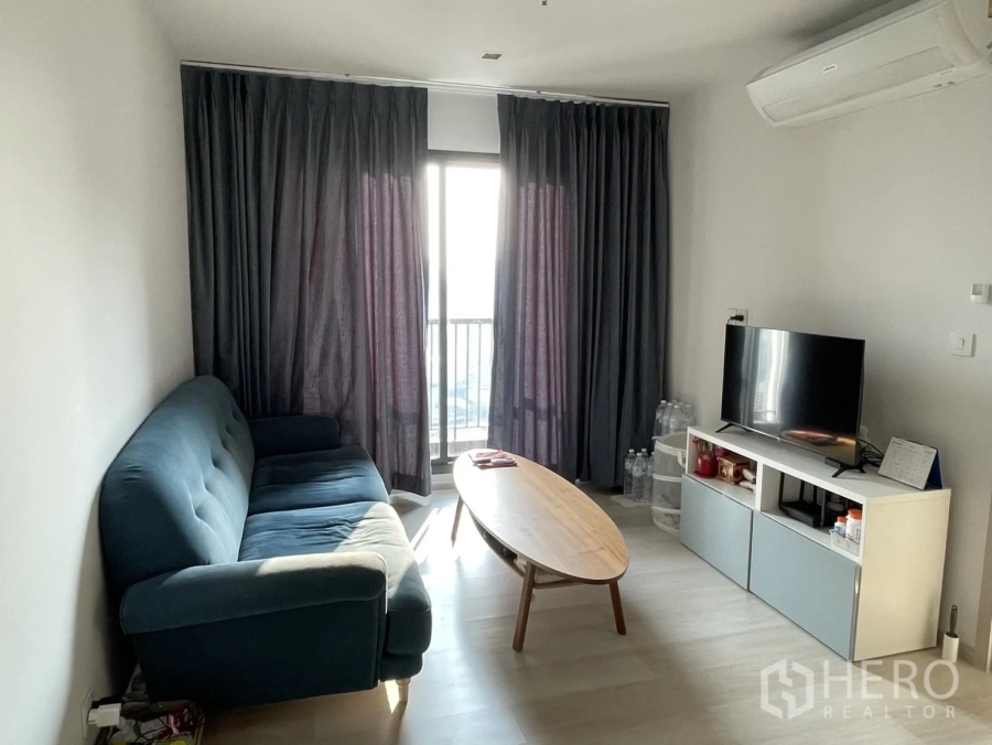 Condominium for rent in Pathum Wan, Bangkok - Living area with blue sofa, TV console and access to a balcony with tall curtains.