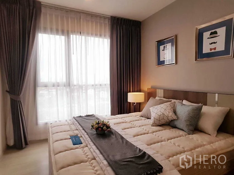 Condominium for sale in Khlong Toei, Bangkok - Cozy bedroom with cushioned bed, bedside lamp, and sheer curtains over a large window.