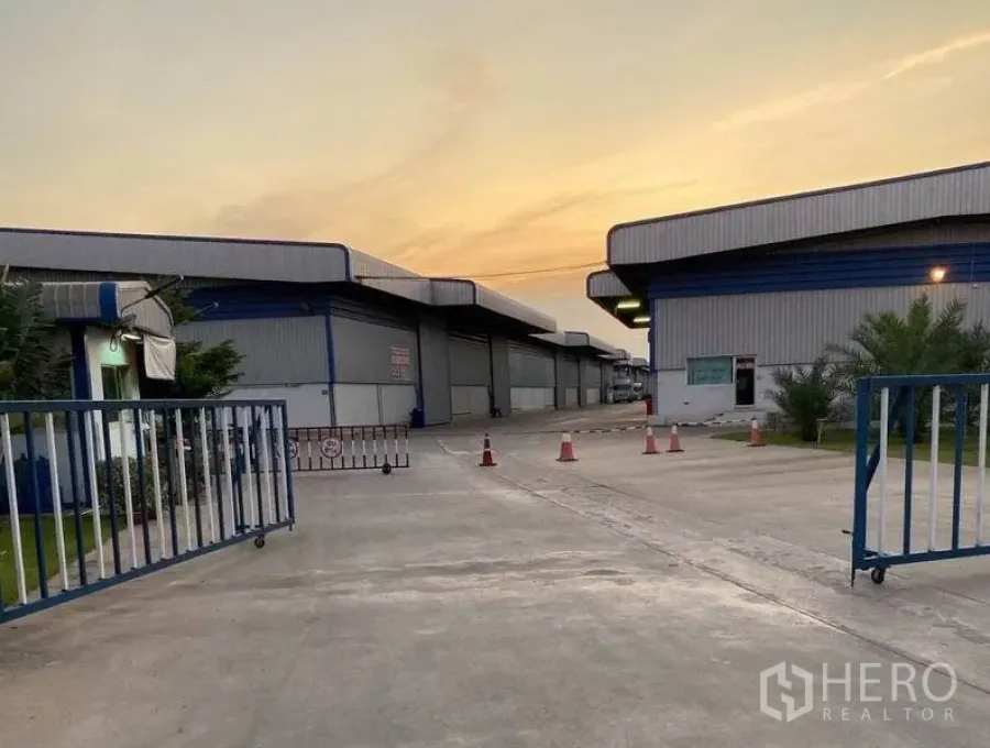 Warehouse for rent in Lat Krabang, Bangkok - Secure gated entrance to a warehouse compound with sliding gate and wide driveway at sunset.