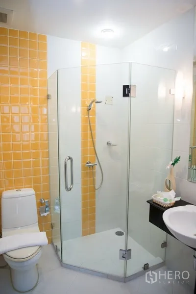 Condominium for rent in Khlong Toei, Bangkok - Modern bathroom with glass shower enclosure, toilet and white vanity beside yellow tiles.