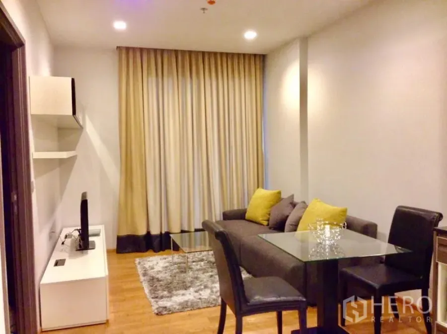 Condominium for rent in Khlong Toei, Bangkok - Cozy living area with gray sofa, glass coffee table, TV unit and floor-to-ceiling curtains.