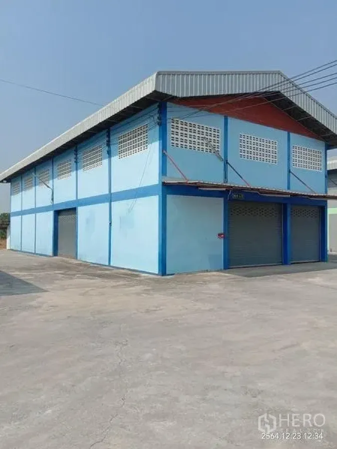 工厂 出租在Lat Lum Kaeo, Pathum Thani - 蓝色厂房转角视图，金属屋顶与多扇卷闸门，前有大院