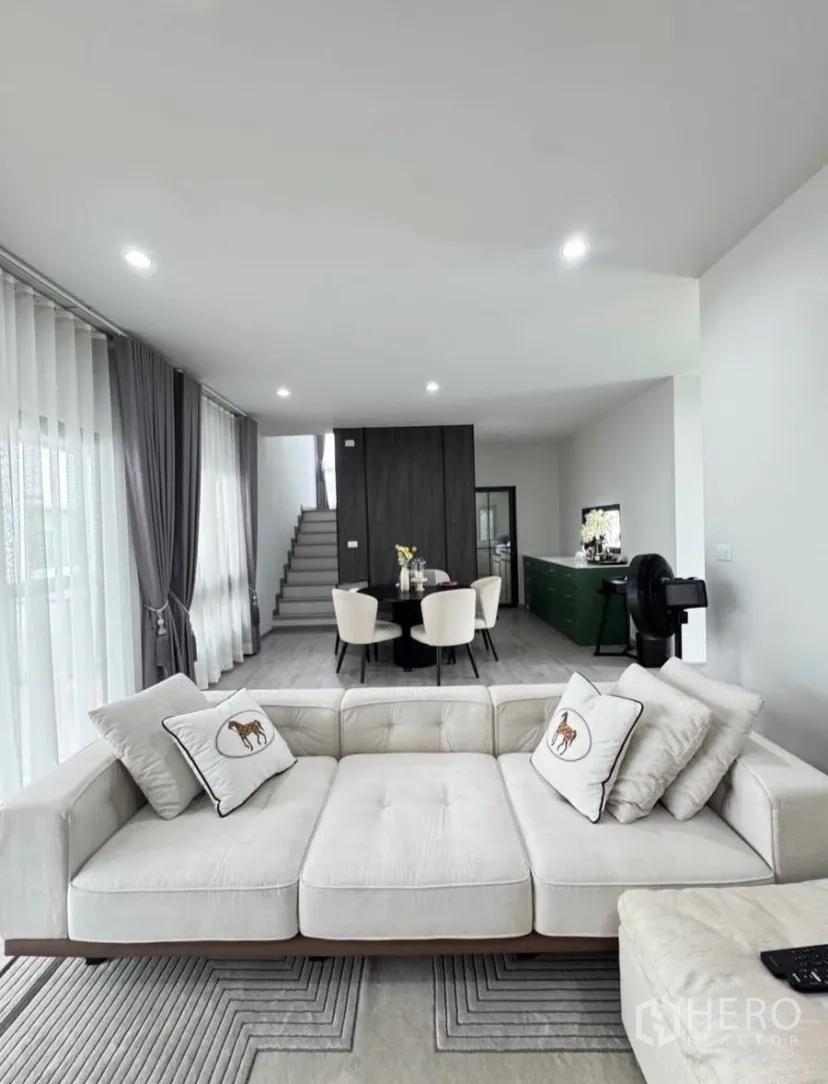 独栋房屋 for rent or sale in Lat Krabang, Bangkok - 开放式客餐厅，大面积落地窗与灰色窗帘，楼梯通向二层。