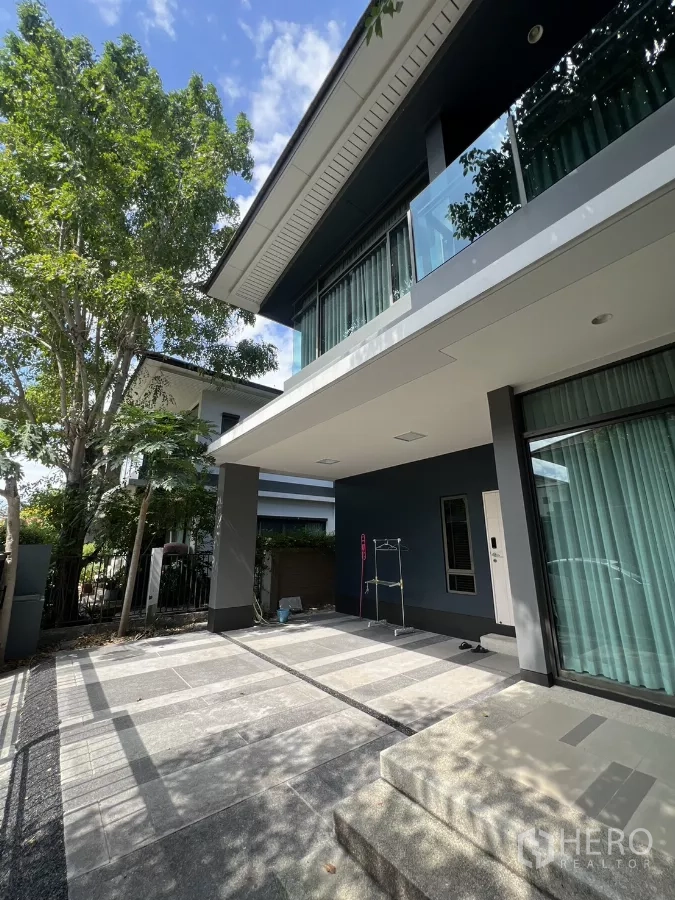 Detached House for rent in Bang Kapi, Bangkok - Covered carport and front patio beneath a cantilevered balcony with greenery around the home.