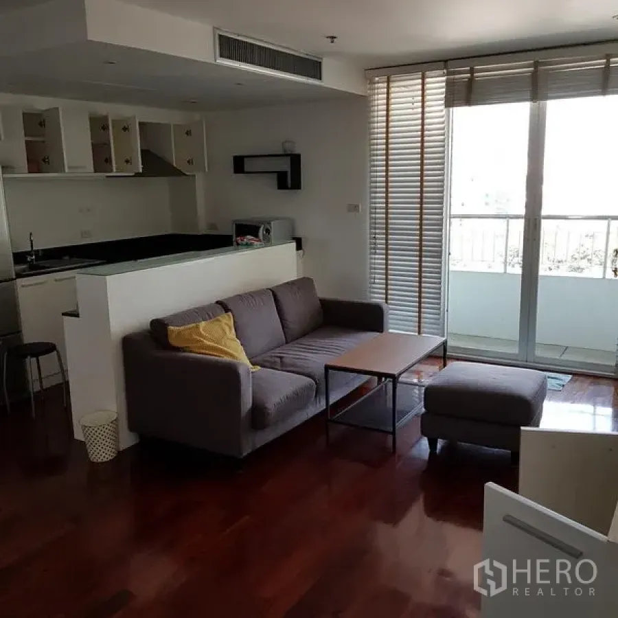 Condominium for rent in Pathum Wan, Bangkok - Bright living area with gray sofa, coffee table, wood floors and sliding glass doors to a balcony beside an open kitchen.