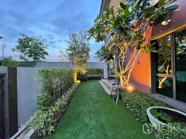Detached House for sale in Pak Kret, Nonthaburi - Landscaped side yard with artificial turf, hedges and a frangipani tree at dusk.