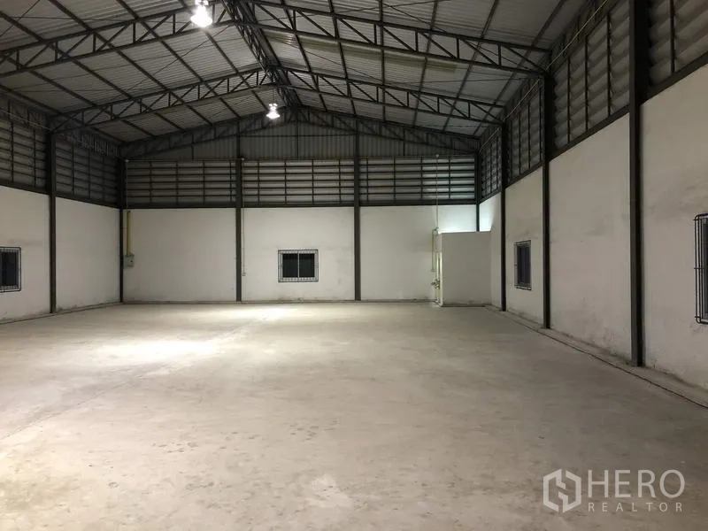 Warehouse for rent in Lam Luk Ka, Pathum Thani - Well‑lit warehouse space at night with concrete floor, high gable roof, and a utility room.