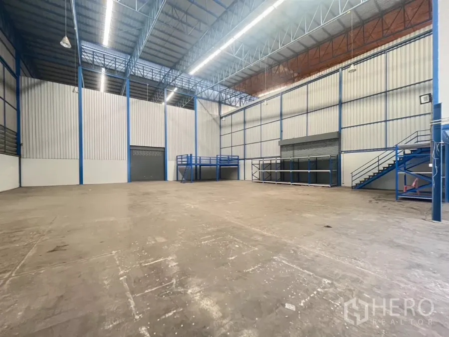 Factory for rent in Phra Pradaeng, Samut Prakan - Spacious warehouse interior with a high steel truss roof, mezzanine platform, roller shutter door and durable concrete floor.