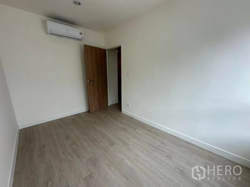 Home Office for rent in Khlong Toei, Bangkok - Compact bedroom with wall-mounted air conditioner, wooden door and neutral walls.