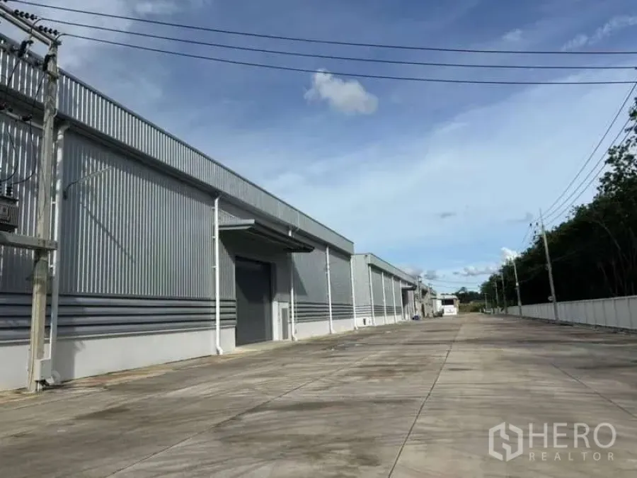 Warehouse for rent in Nikhom Phatthana, Rayong - Long service road beside corrugated metal warehouse buildings with power lines