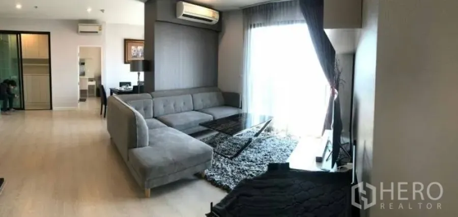 Condominium for rent or sale in Huai Khwang, Bangkok - Bright living room with L-shaped sofa, shag rug and balcony window in a modern condo.