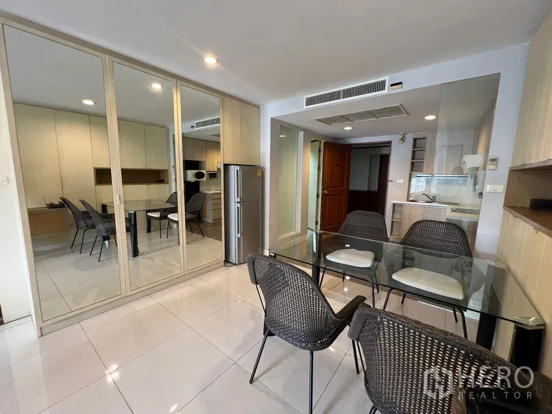 Condominium for rent or sale in Khlong Toei, Bangkok - Dining area with glass-top table, mirrored closets and a kitchenette beyond.
