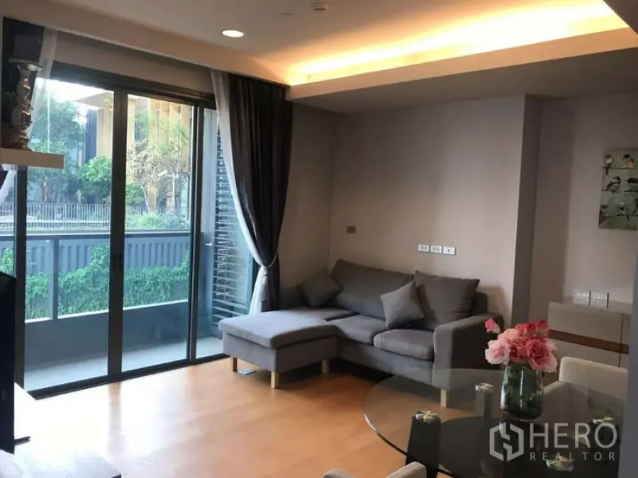 Condominium for rent in Khlong Toei, Bangkok - Bright living room with gray L-shaped sofa, wood floors and sliding doors to a balcony with garden view.