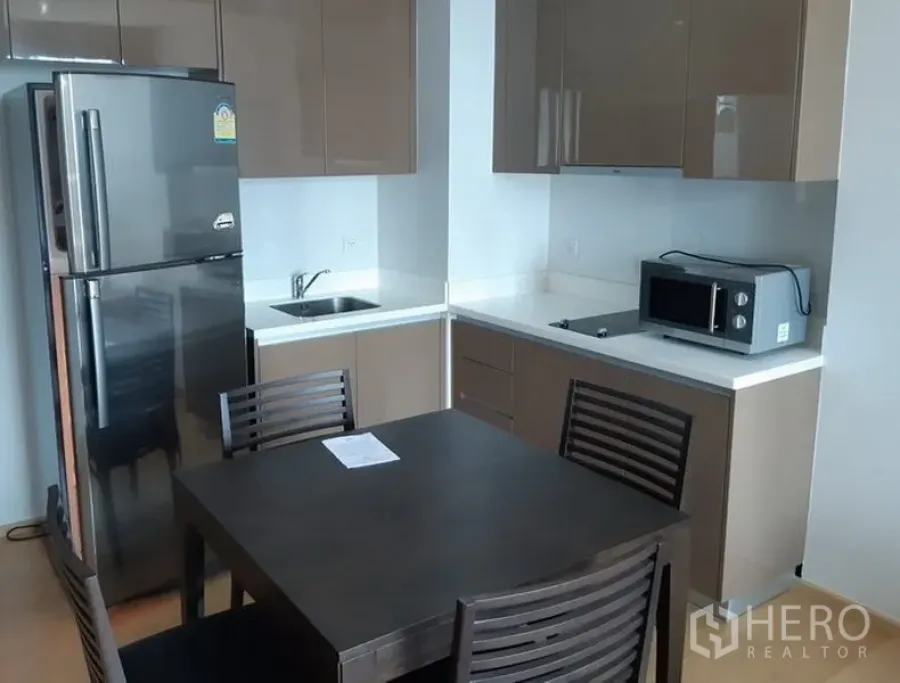 Condominium for rent in Khlong Toei, Bangkok - Modern L-shaped kitchen in a Khlong Toei, Bangkok condo with glossy cabinets, stainless fridge, microwave and a four-seat dining table.