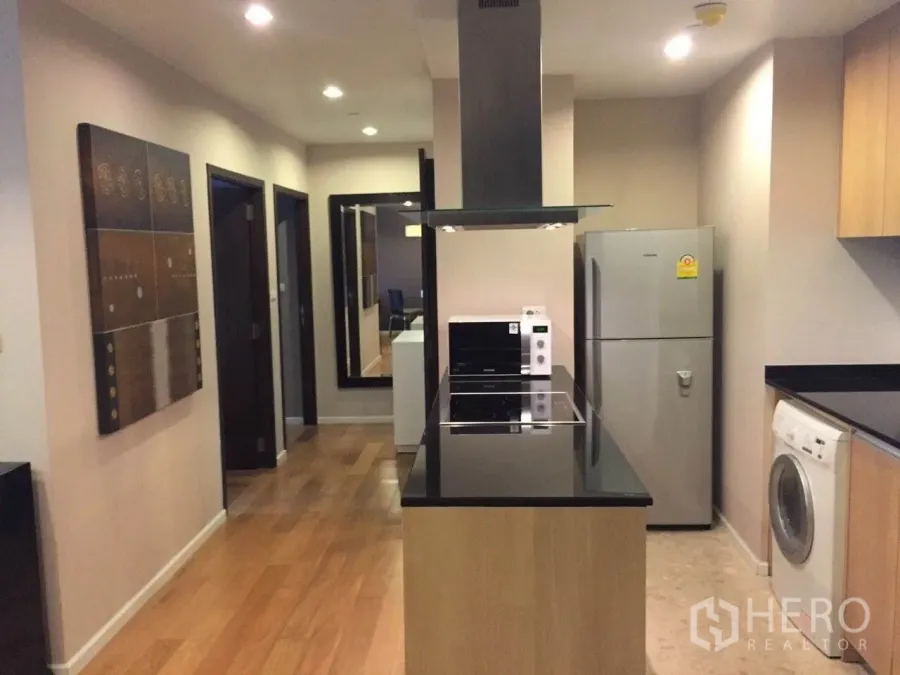 Condominium for rent or sale in Sathon, Bangkok - Modern kitchen with black granite island, cooktop, hood, fridge and in‑unit washer beside a wood‑floored hallway.