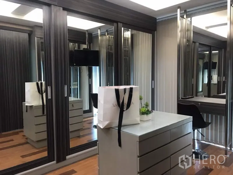 Townhouse for rent or sale in Suan Luang, Bangkok - Walk-in closet with mirrored wardrobes, central drawer island, vanity desk, and wooden flooring.