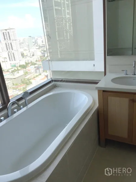 Condominium for rent in Khlong Toei, Bangkok - Bathroom with a soaking bathtub beside a large window offering city skyline views and a vanity sink.