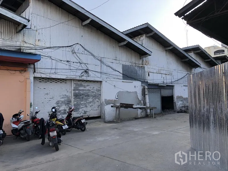 Retail Space for rent in Khlong Toei, Bangkok - Corrugated metal warehouse exterior with multiple roller shutter doors and an open loading forecourt with parked motorbikes.