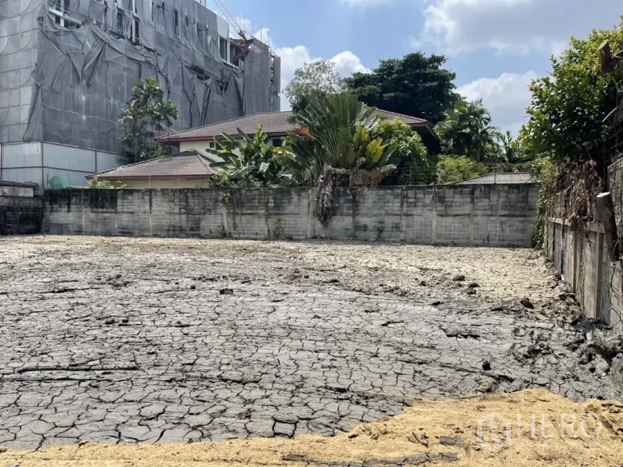 Land for sale in Phra Khanong, Bangkok - Walled vacant land in Phra Khanong near BTS Punnawithi, cracked soil with neighboring houses, trees, and a covered building in the background.