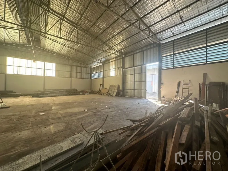 Warehouse for rent in Saphan Sung, Bangkok - Wide warehouse space showing large sliding steel door, ventilated louver panels and scattered lumber.