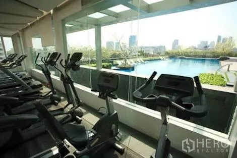 Condominium for rent in Khlong Toei, Bangkok - Condominium fitness center with cardio machines overlooking an outdoor pool and city skyline.