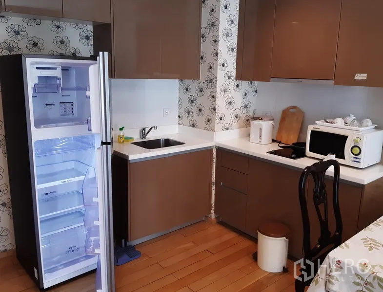 Condominium for rent in Khlong Toei, Bangkok - Modern kitchenette with brown cabinets, white countertops, open refrigerator, microwave and electric cooktop.