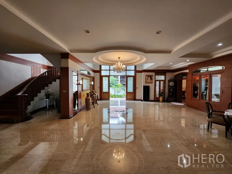Detached House for rent in Prawet, Bangkok - Expansive living area with chandelier, high windows and a sweeping wooden staircase to the upper floor.