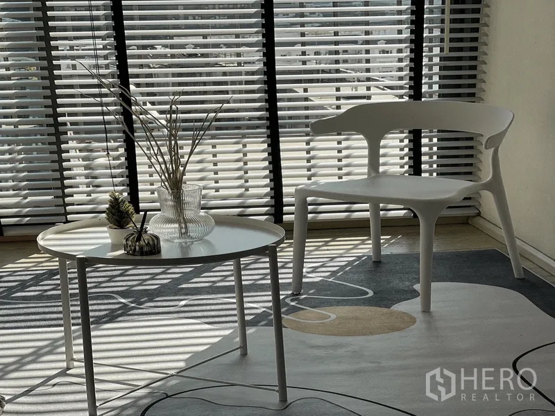 Condominium for rent in Khlong Toei, Bangkok - Close‑up of round coffee table and modern white chair beside window blinds and patterned rug.