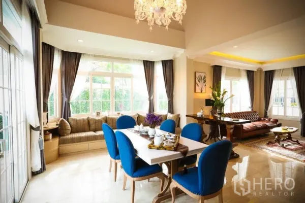 Detached House for rent in Bang Na, Bangkok - Bright open-plan living and dining room with chandelier, large windows, sofa set and blue dining chairs.