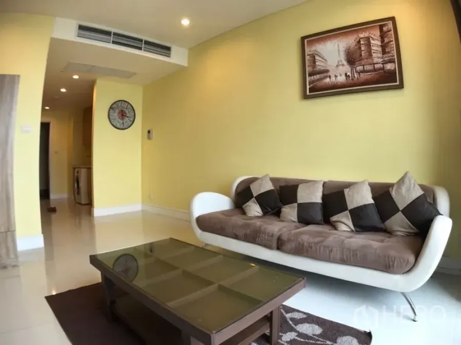 Condominium for rent in Khlong Toei, Bangkok - Bright living room with yellow walls, modern sofa, glass coffee table and wall art leading to a hallway with recessed lighting.