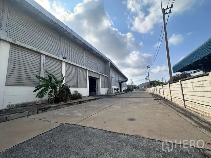Factory for rent - Exterior of a large industrial warehouse with roller shutters and a wide loading driveway under a cloudy sky.