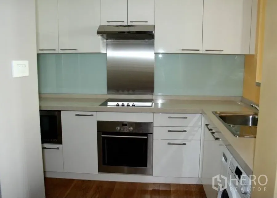 Condominium for rent or sale in Watthana, Bangkok - Modern L-shaped kitchen with white cabinetry, glass backsplash, built-in oven and cooktop, and wood flooring in a Bangkok condo.