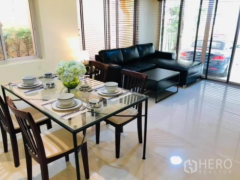 Detached House for rent in Watthana, Bangkok - Bright dining and living area with a glass dining table and black leather sectional beside large windows.