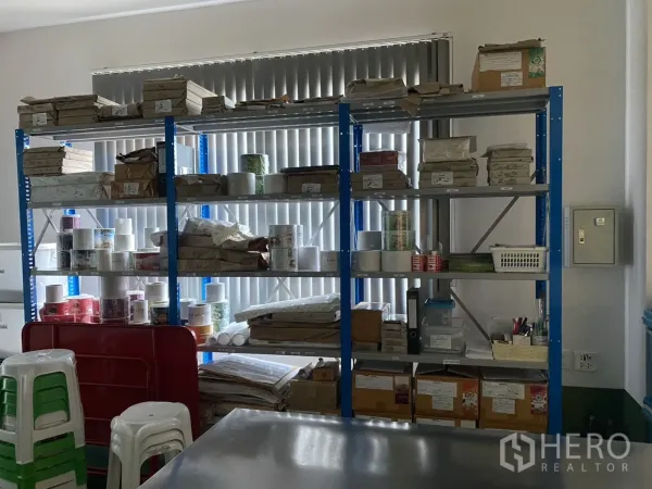 Factory for sale in Khlong Luang, Pathum Thani - Storage room with blue racks holding supplies beside vertical window blinds.