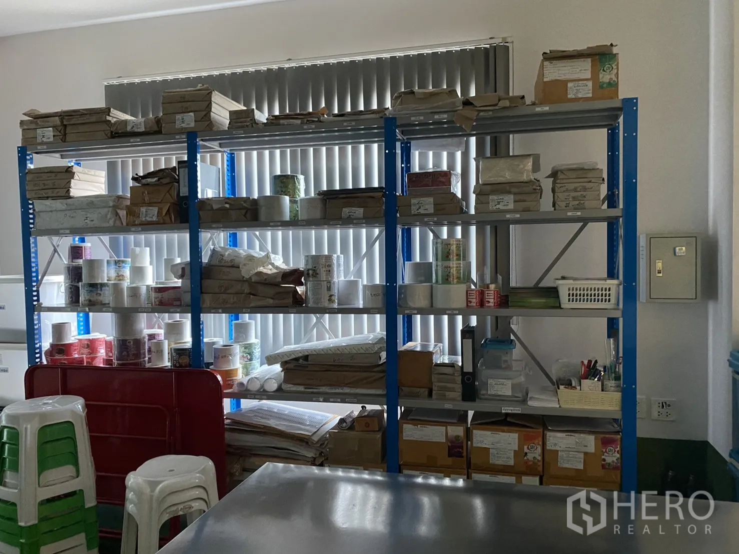 Factory for sale in Khlong Luang, Pathum Thani - Storage room with blue racks holding supplies beside vertical window blinds.