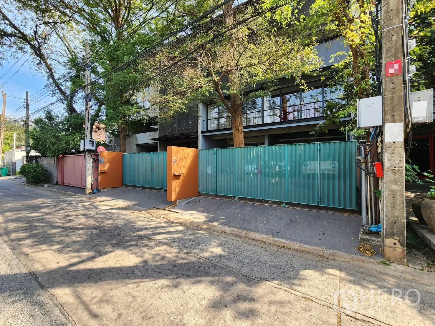 Home Office for rent in Chatuchak, Bangkok - Street view of gated entrance with colored panels and mature trees.