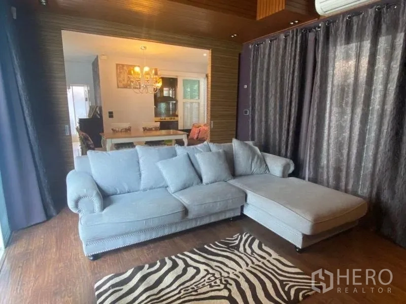 Detached House for rent or sale in Prawet, Bangkok - Cozy family lounge with L-shaped sofa, zebra rug, and dark curtains facing dining area.