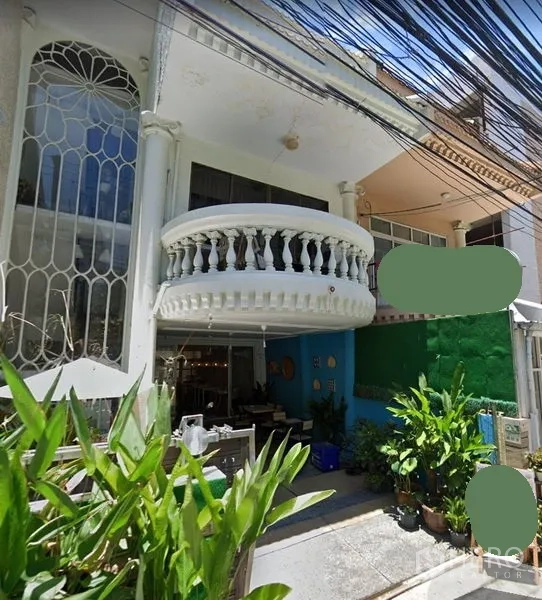 Townhouse for rent in Watthana, Bangkok - Street-level view of a white Bangkok townhouse with a curved balcony, tall arched window, leafy front patio, and overhead cables in daylight.