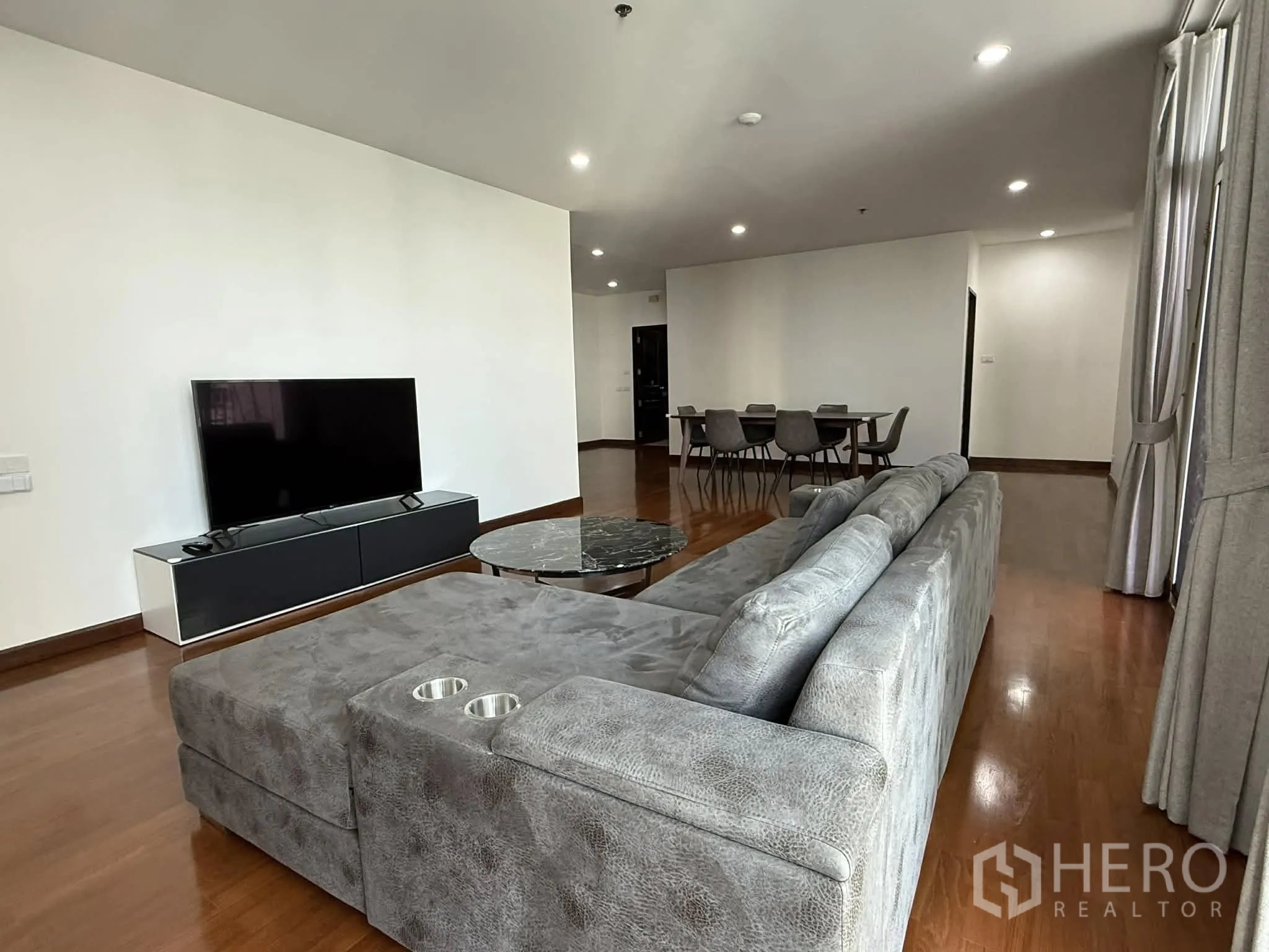 Condominium for rent in Pathum Wan, Bangkok - Living room with a large gray sectional sofa, TV console, and round marble table.