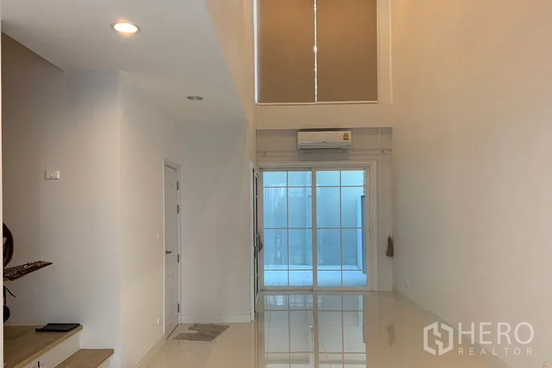 Townhouse for rent in Bang Na, Bangkok - Double-height living room with glossy tile floor, AC and a tall window above sliding glass doors