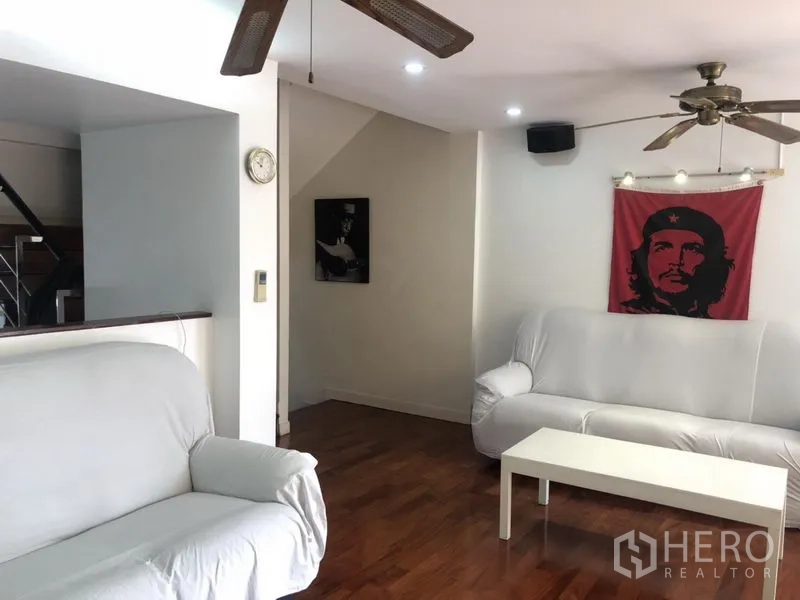 Townhouse for rent or sale in Bangkok - Cozy living room with white sofas, wood flooring and a ceiling fan, accented by wall art.