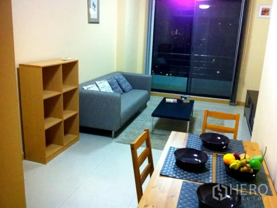 Condominium for rent or sale in Watthana, Bangkok - Bright living and dining area with grey sofa, shelving, coffee table, and sliding glass doors to a balcony with city night views.