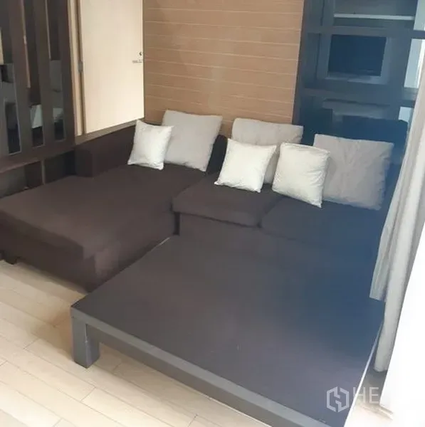 Condominium for rent in Khlong Toei, Bangkok - Spacious living area featuring a dark L-shaped sofa and neutral wood floors.