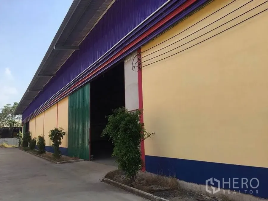คลังสินค้า ให้เช่าในSai Noi, Nonthaburi - ด้านหน้าโกดัง ผนังสีเหลือง หลังคาสีม่วง ประตูสไลด์สีเขียว และทางคอนกรีต