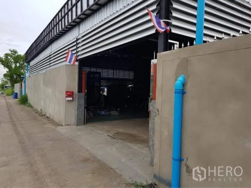 Warehouse for sale in Lam Luk Ka, Pathum Thani - Full exterior of metal-sheet warehouse behind a high wall along a quiet lane.