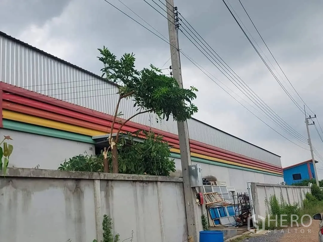 Warehouse for sale in Lam Luk Ka, Pathum Thani - Side exterior of the warehouse behind a concrete wall with ventilation panels and overhead utility lines.