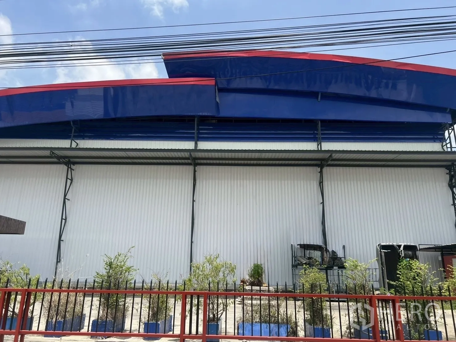 Warehouse for rent in Lat Krabang, Bangkok - Front elevation of a Lat Krabang warehouse with blue-and-red roof fascia, white steel cladding and a fenced landscaped verge.